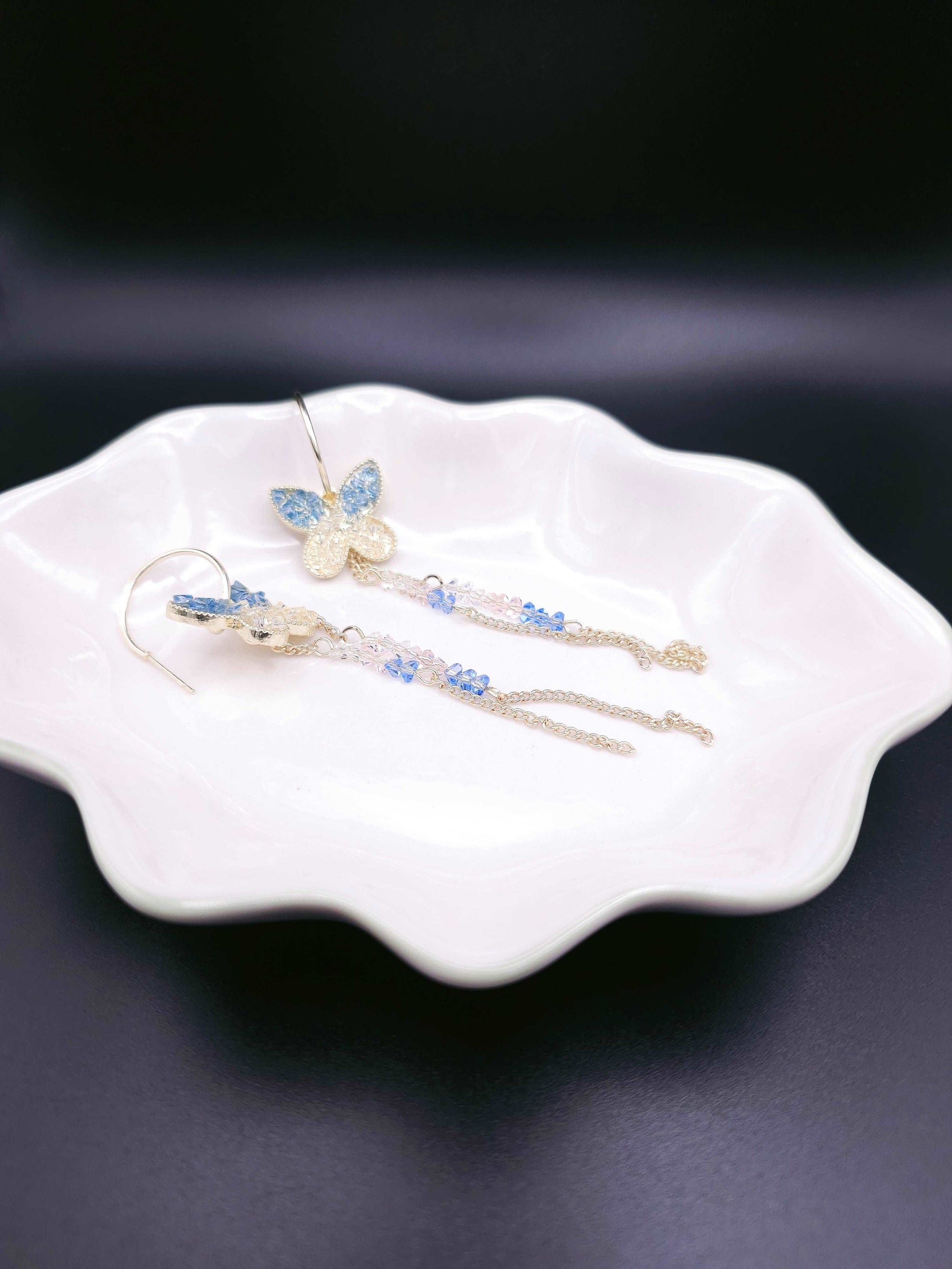 Lay down shot of Fairy-tale Earrings displaying the stunning butterfly dangle design, gold base, and blue and clear sparkles in an elegant arrangement.